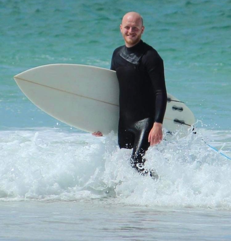 Surf and paddle board coach Lewis (Boris) walking out of a turquiose sea with a surf board