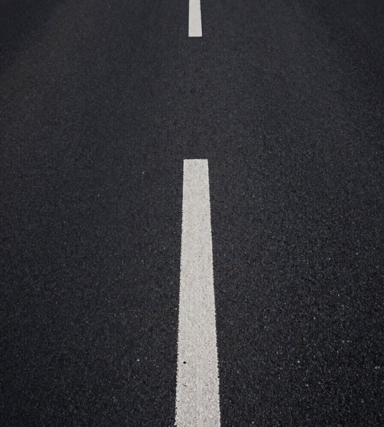 Extreme close-up overhead view of a clean asphalt road stretching into the distance, a single white dashed center line cutting through deep shadow and pale light, minimal and geometric, abstract road geometry with no people, high contrast studio-quality lighting, wide negative space on the left side