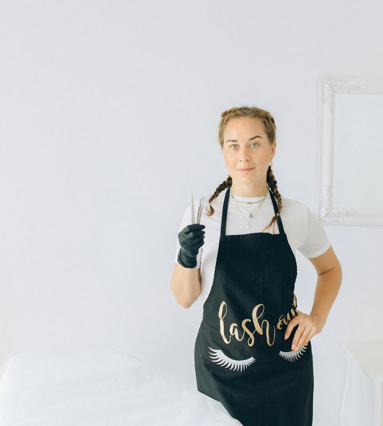 Artista de pestañas profesional con un delantal negro sosteniendo pinzas en un luminoso estudio