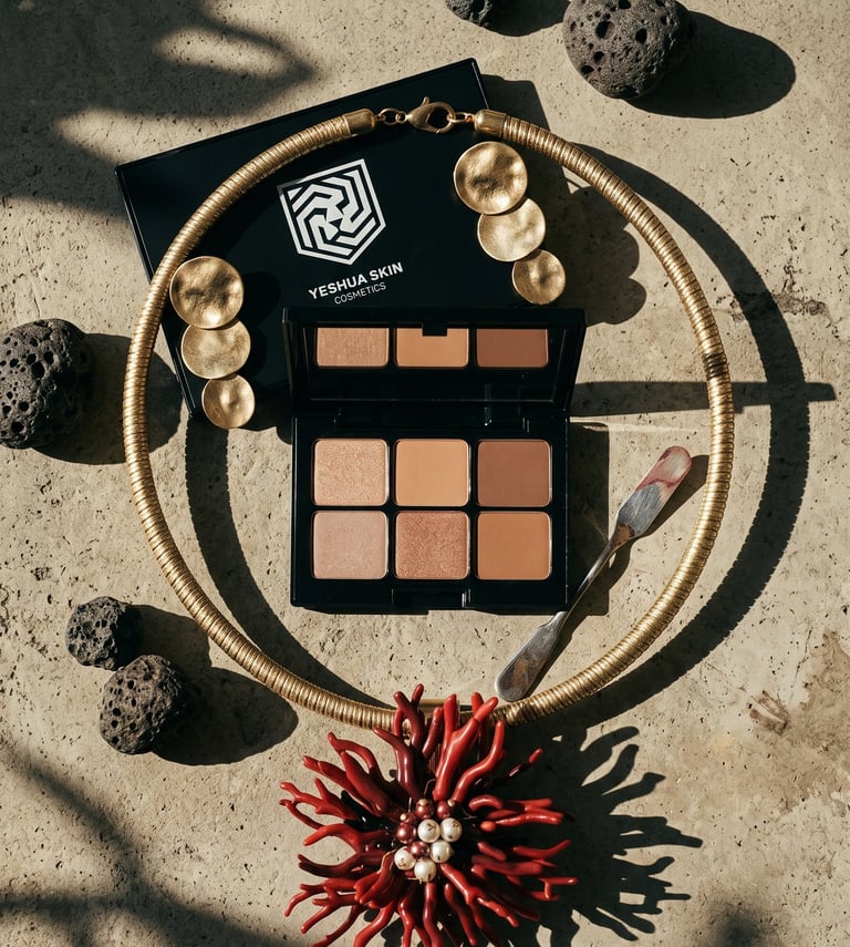 Yeshua Skin Cosmetics eyeshadow palette with gold jewelry and volcanic rocks on a stone surface.
