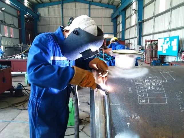 Professional welder in protective gear welding a large industrial steel pipe in a fabrication workshop.