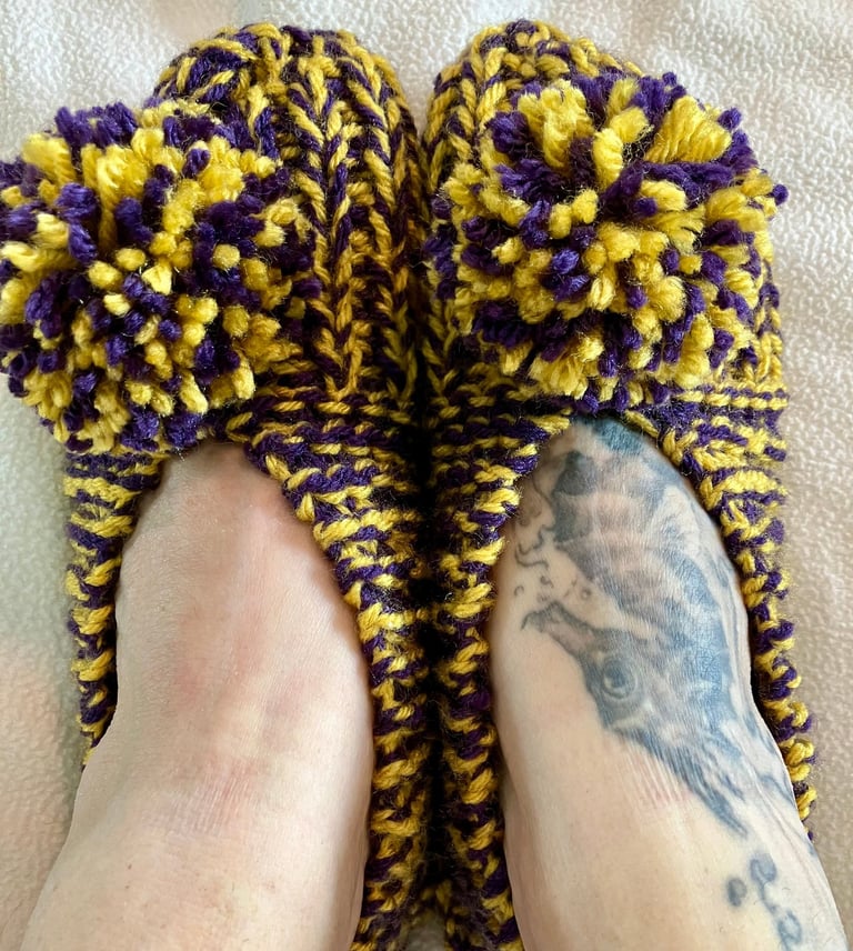 Close up of purple and yellow hand knit house shoes with pompoms on models feet