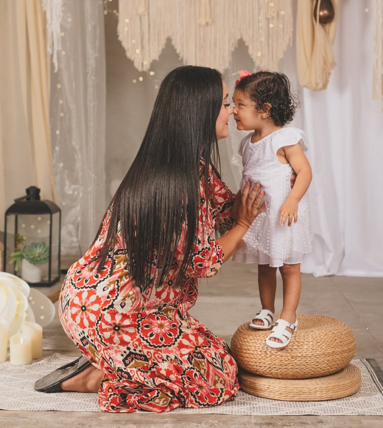 una mamá hincada y su niña pequeña parada, tocándose nariz con nariz
