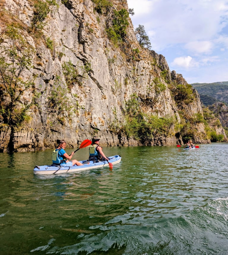 Tourists kayaking in blue boats along a steep Matka canyon wall on green river water.