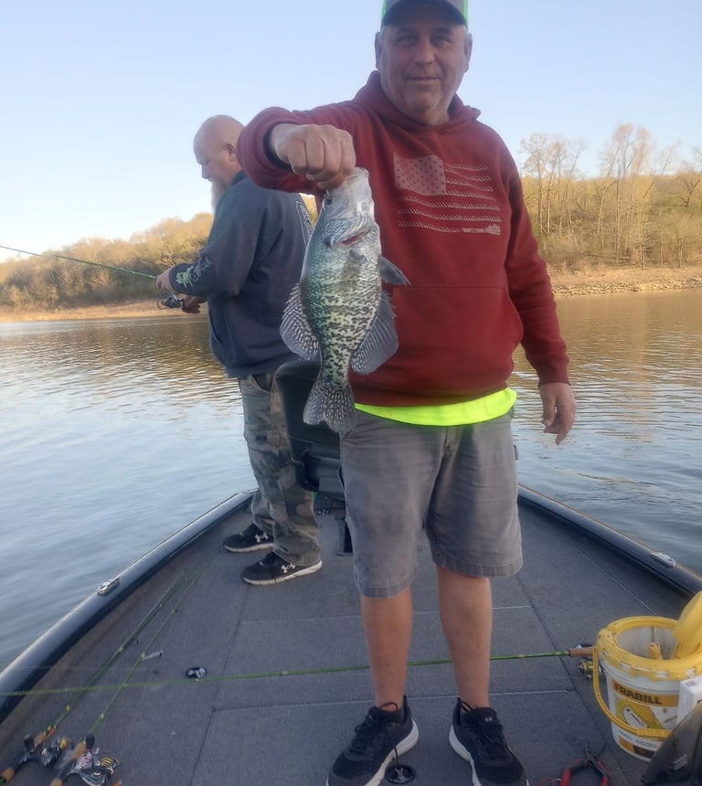 A smiling man on a fishing boat holding up a freshly caught crappie fish on a sunny lake.