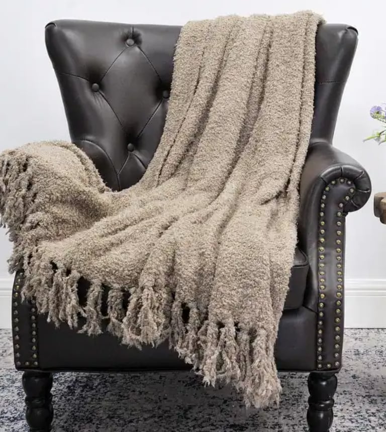 Cozy taupe textured throw blanket with tassels draped over a dark leather tufted armchair.