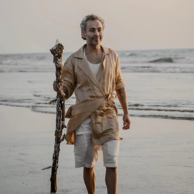 A man in linen clothing walks on a sandy beach holding a decorative wooden staff at sunset.