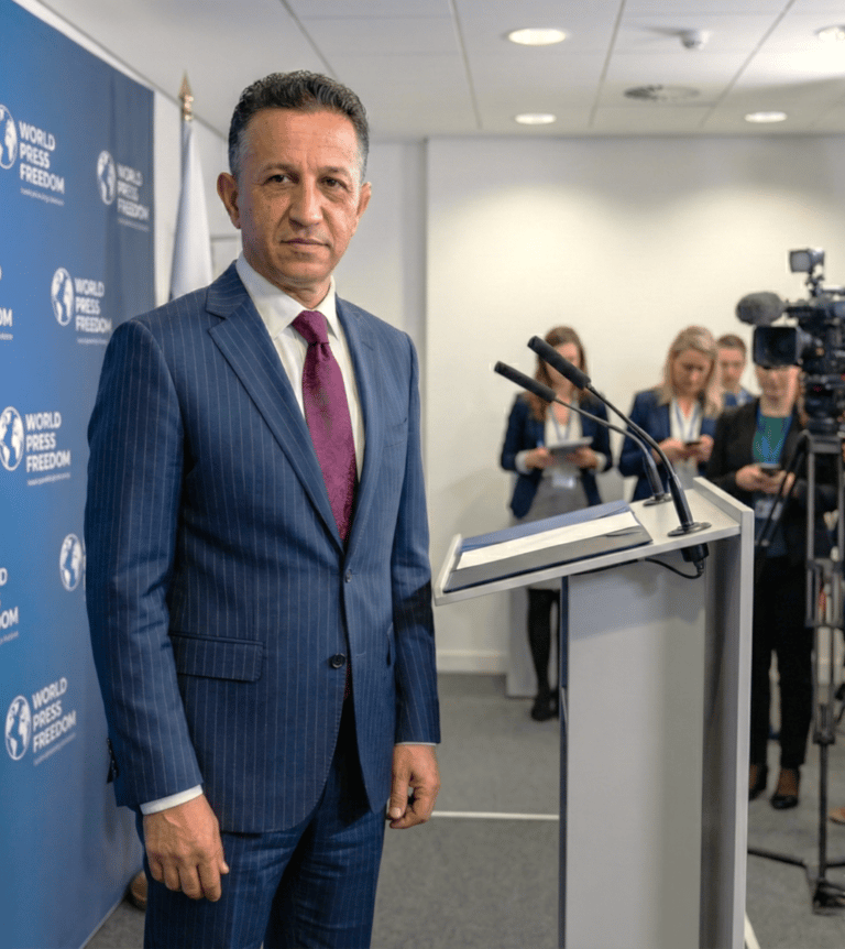 A speaker in a suit delivering remarks at an international press event, standing behind a podium. 