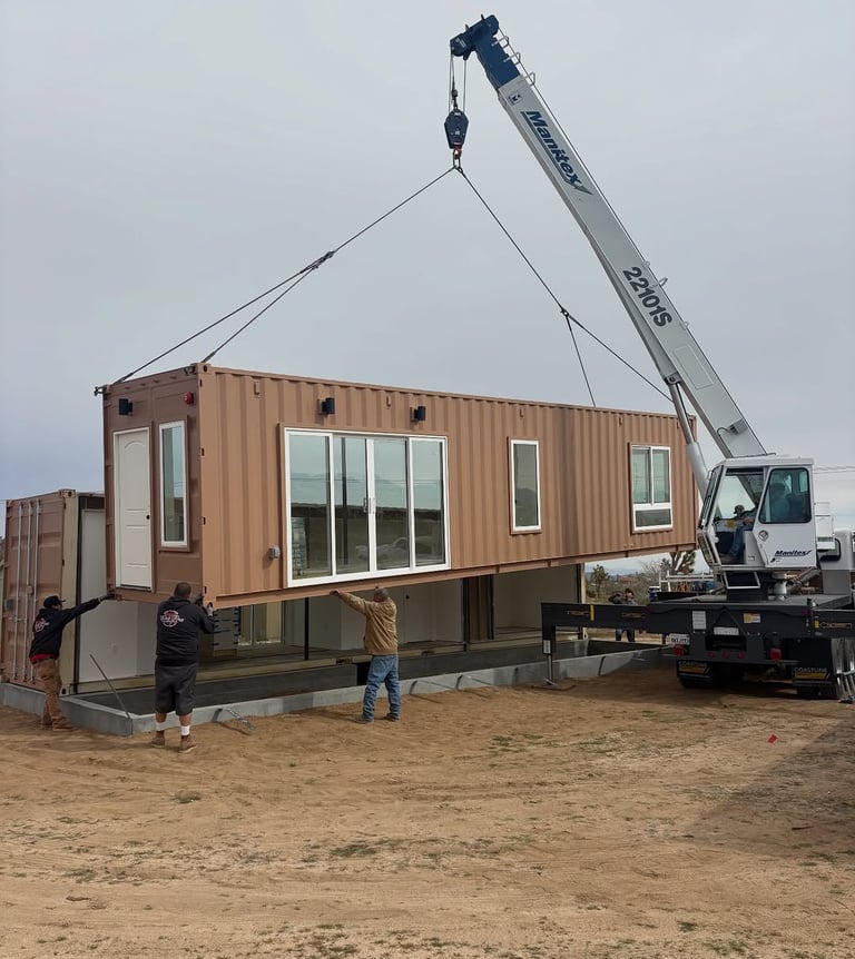 A construction crane lifts a modular shipping container home onto a foundation at a build site.