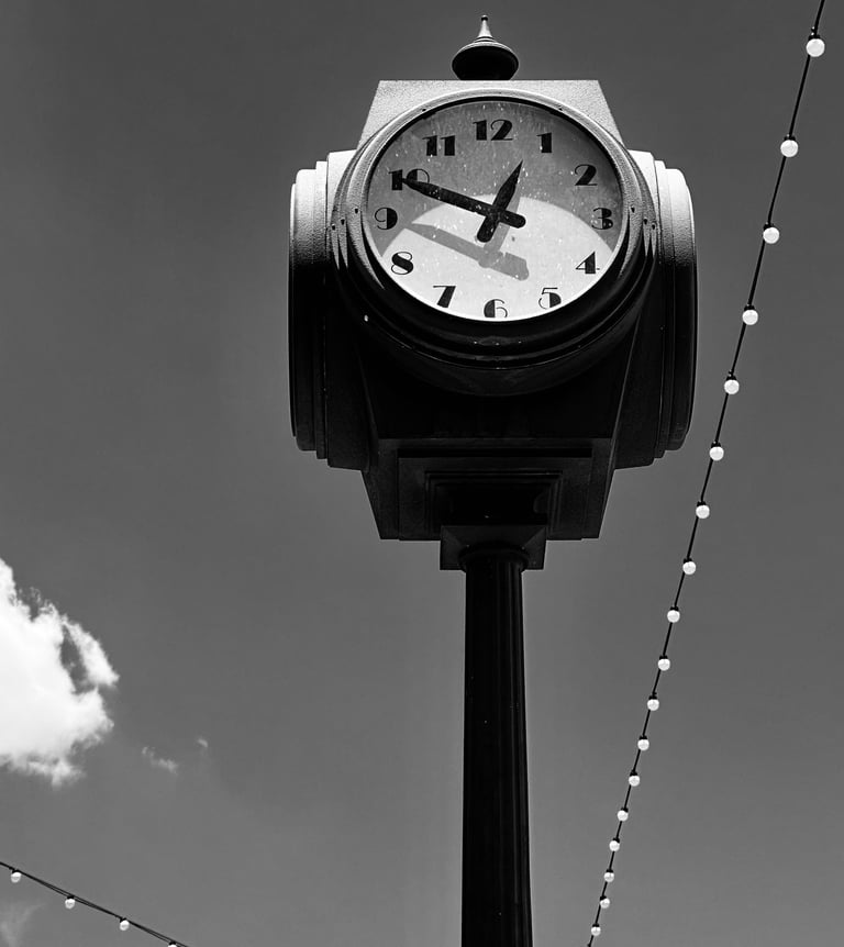 A black-and-white photograph of a vintage street clock mounted on a tall post against a clear sky.