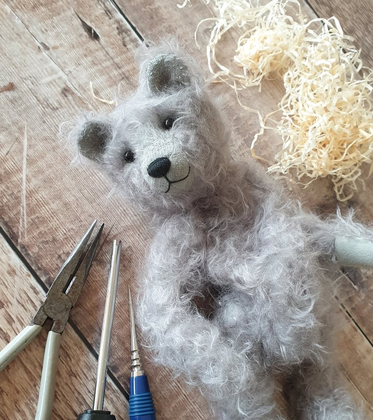 a partly made handmade teddy bear in grey mohair with a wistful face and surrounded by tools on a table