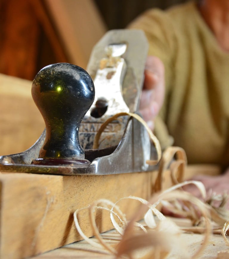 A woodworker uses a vintage metal hand plane to smooth a wooden board with curly shavings.