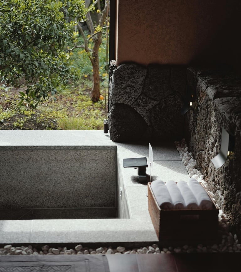Luxury indoor-outdoor stone soaking tub with rolled white towels overlooking a lush green garden.