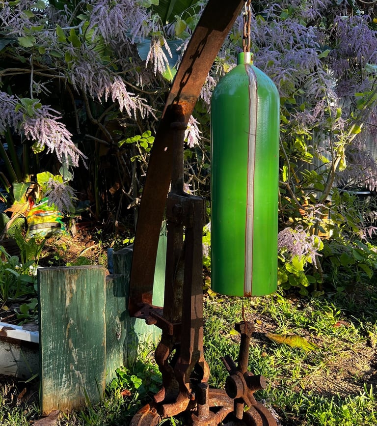 Rustic garden wind chime made from a repurposed green oxygen tank hanging from antique metal farm machinery.