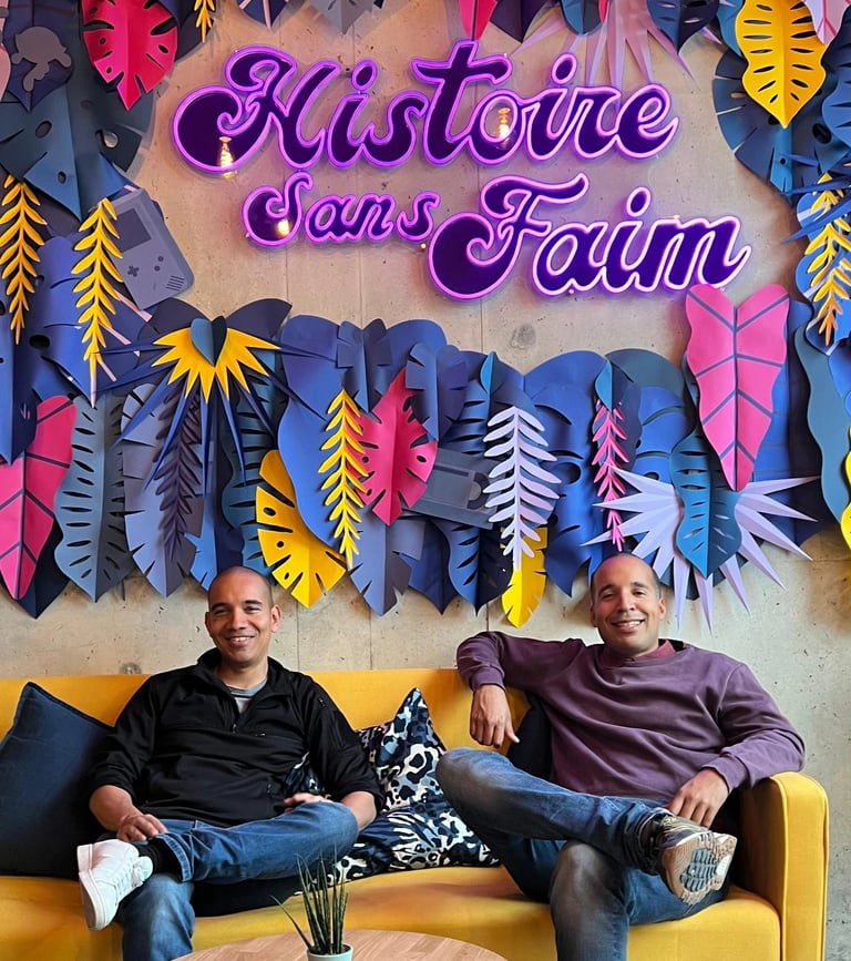 Christopher et Michael assis dans un canapé jaune sous le néon violet Histoire Sans Faim, décor pop coloré.