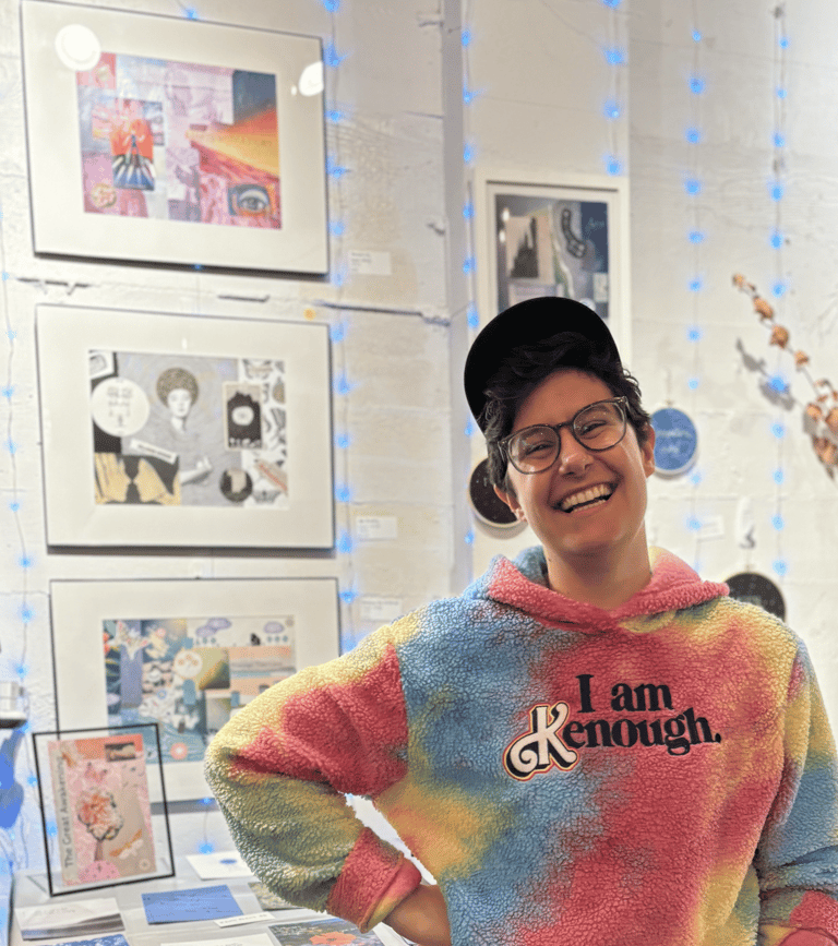 Artist Wren laughs at the camera in front of their art booth displaying their work.