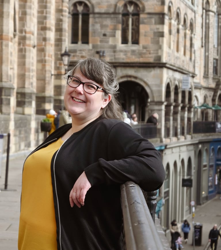 Laura is leaning against a railing with a soft smile on her face, turned towards the camera