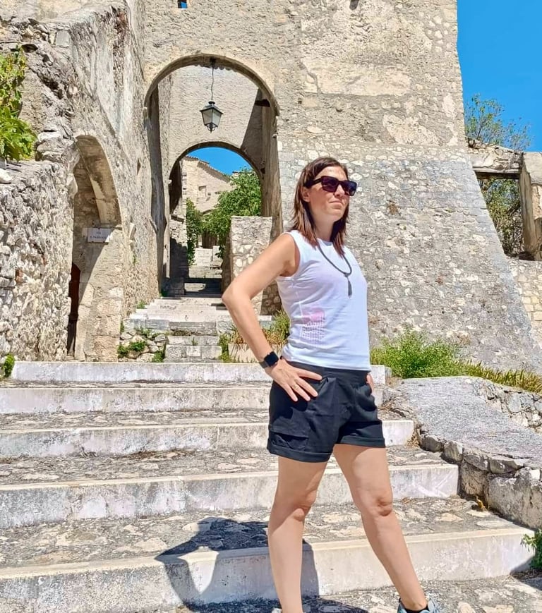 A female traveler in sunglasses stands on stone steps at an ancient European fortress gateway.