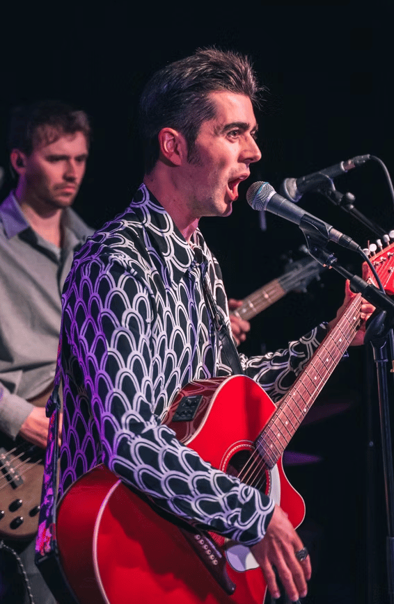 Live performance in Brighton featuring Olly Williams singing and playing guitar on stage with colourful lighting.