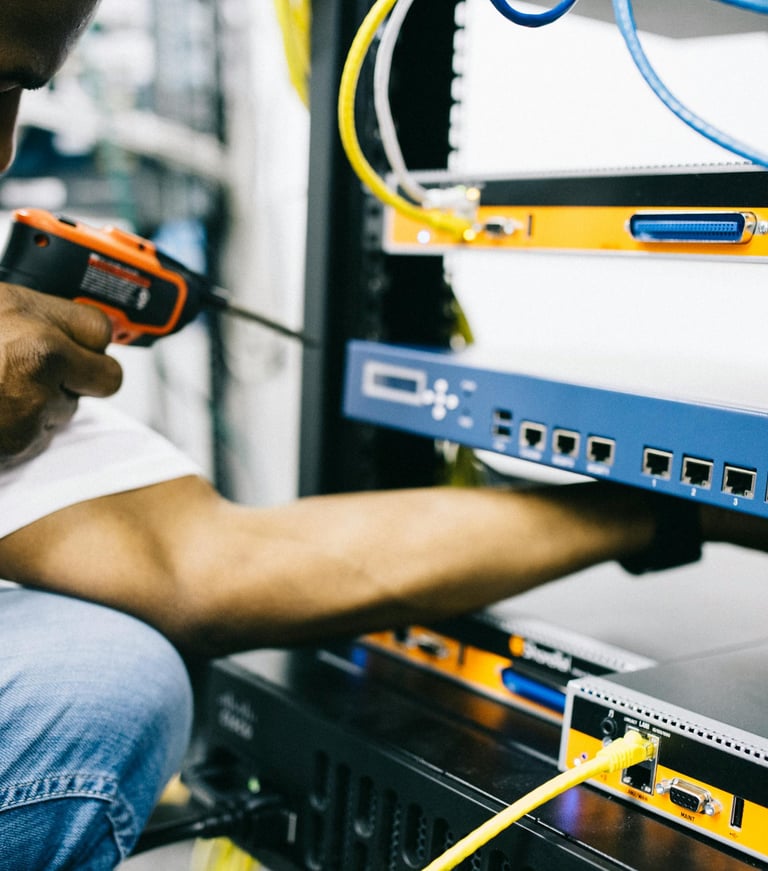 A close-up of a person working on a server rack, connecting network cables with a drill in hand and visible Ethernet ports.