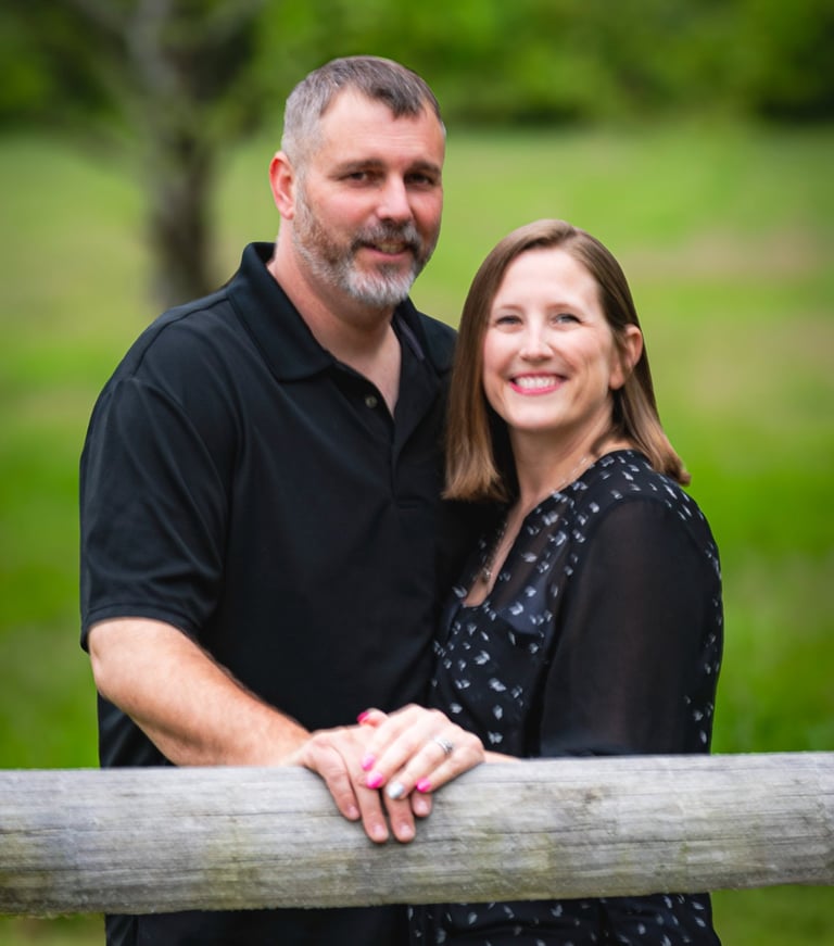 A smiling couple posing for an outdoor portrait by a rustic wooden fence in a lush green field.