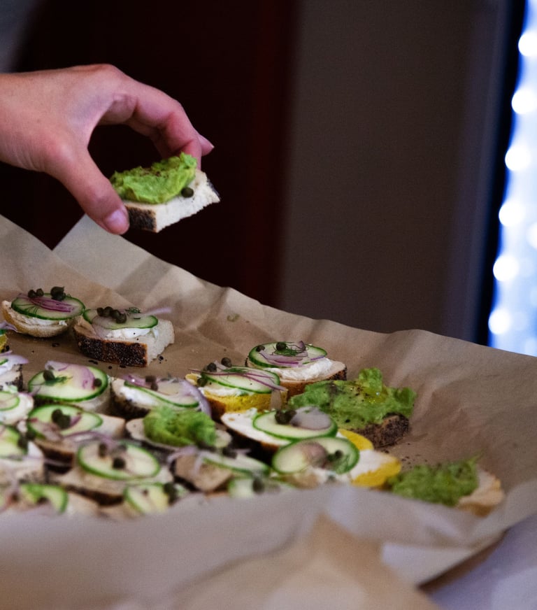 A hand picking up a small piece of bread with avocado on top from a large tray of similar snacks