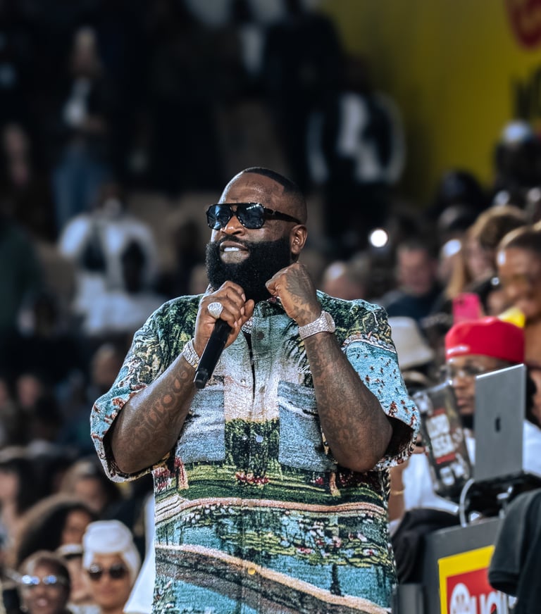 Rapper Rick Ross performing on stage in a scenic tapestry shirt and sunglasses with a microphone.