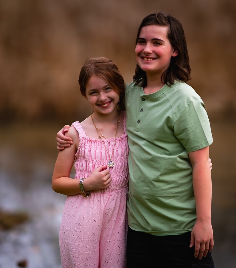 Smiling young siblings posing together outdoors with the older boy's arm around his younger sister. 