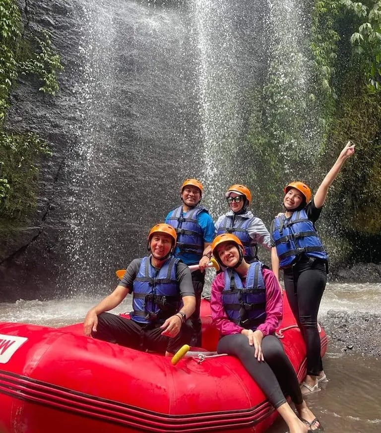 Happy group in life jackets and helmets posing on a red whitewater rafting boat near a waterfall.