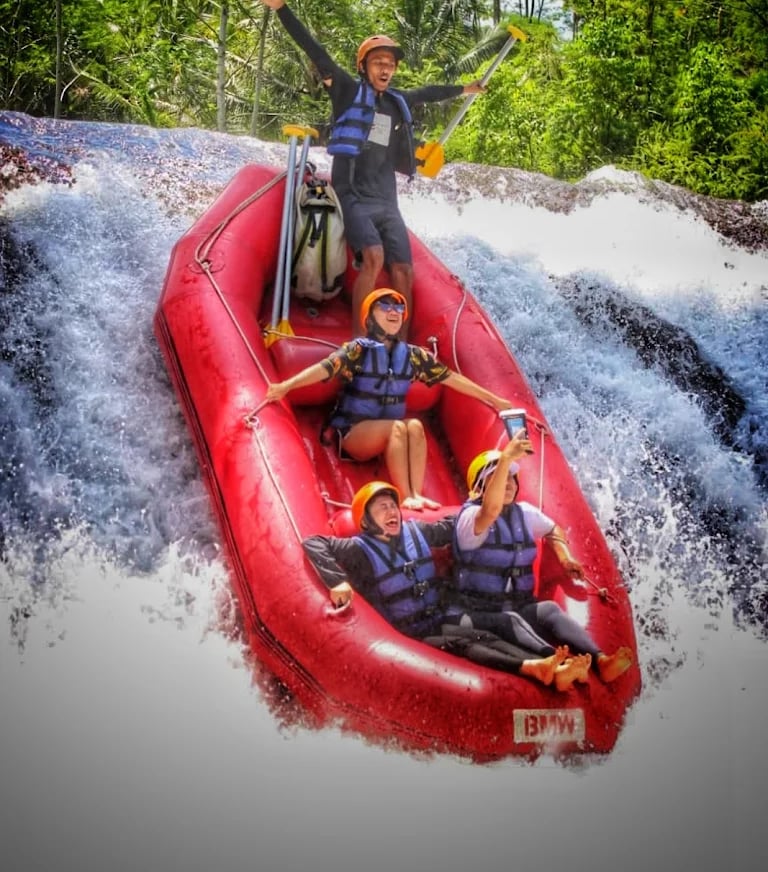 A group of tourists in a red inflatable boat white water rafting down a waterfall in a tropical jungle.