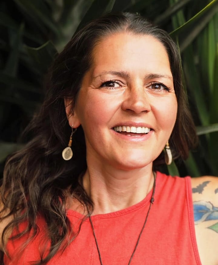 Smiling woman with brown hair and red top posing in front of lush green tropical plants.