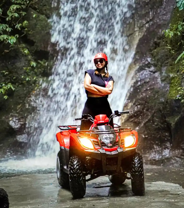 A smiling woman in a red helmet posing on a red ATV quad bike in front of a tropical jungle waterfall.