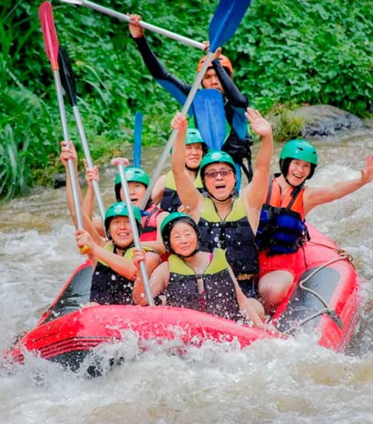 A group of happy people in helmets white water rafting down a river with paddles raised.