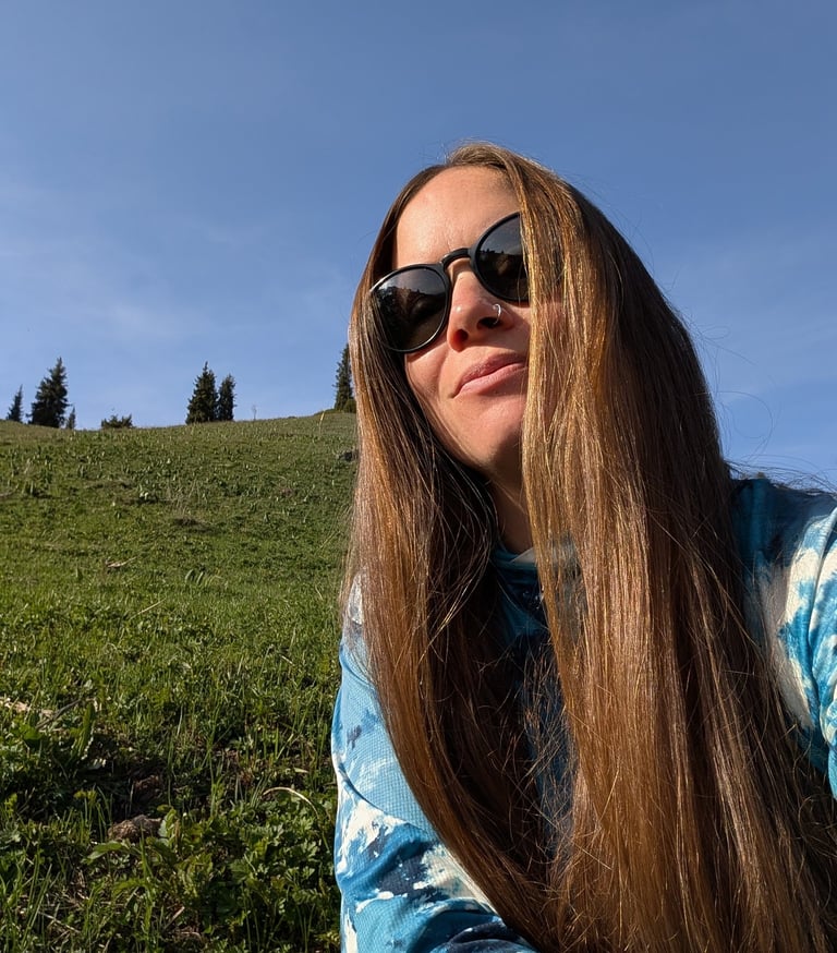 A long-haired woman wearing sunglasses enjoys a sunny day on a grassy green hillside under a clear blue sky.