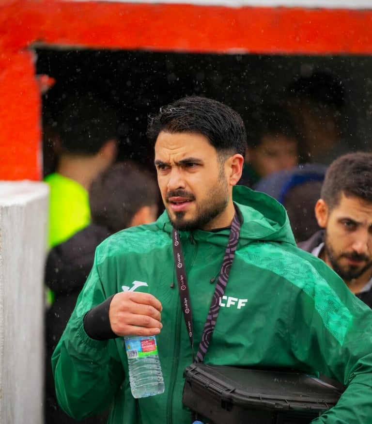A soccer coach in a green jacket holding a water bottle at a football stadium during a game.