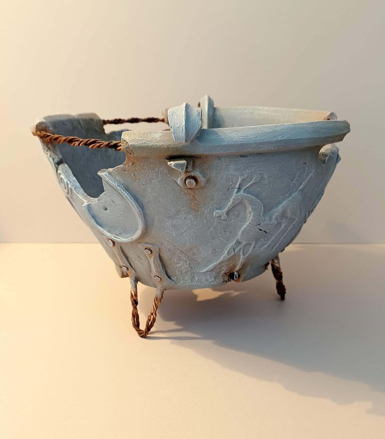 A fragmented, round bowl-like sculpture adorned with symbols and letters, finished in blue-grey hues