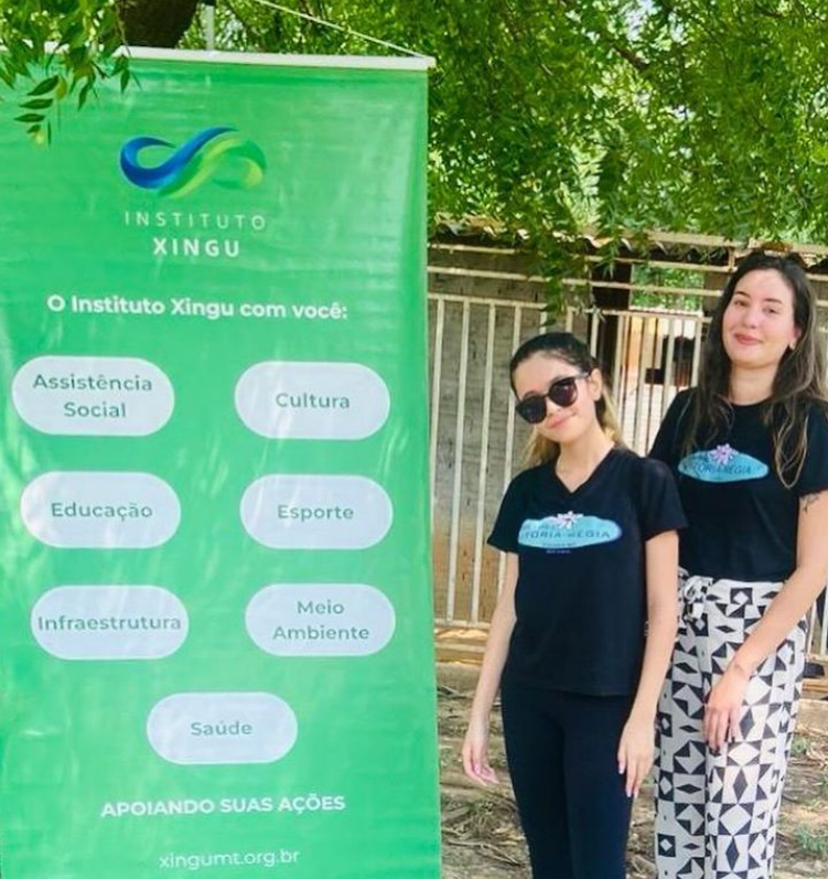 Uma criança e uma mulher em pé ao lado de um banner do Instituto Xingu MT