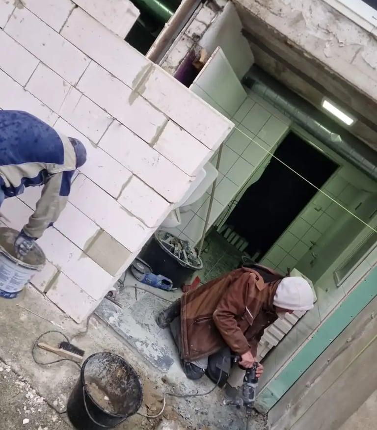 Construction workers building a white aerated concrete block wall and using a power tool on a renovation site.