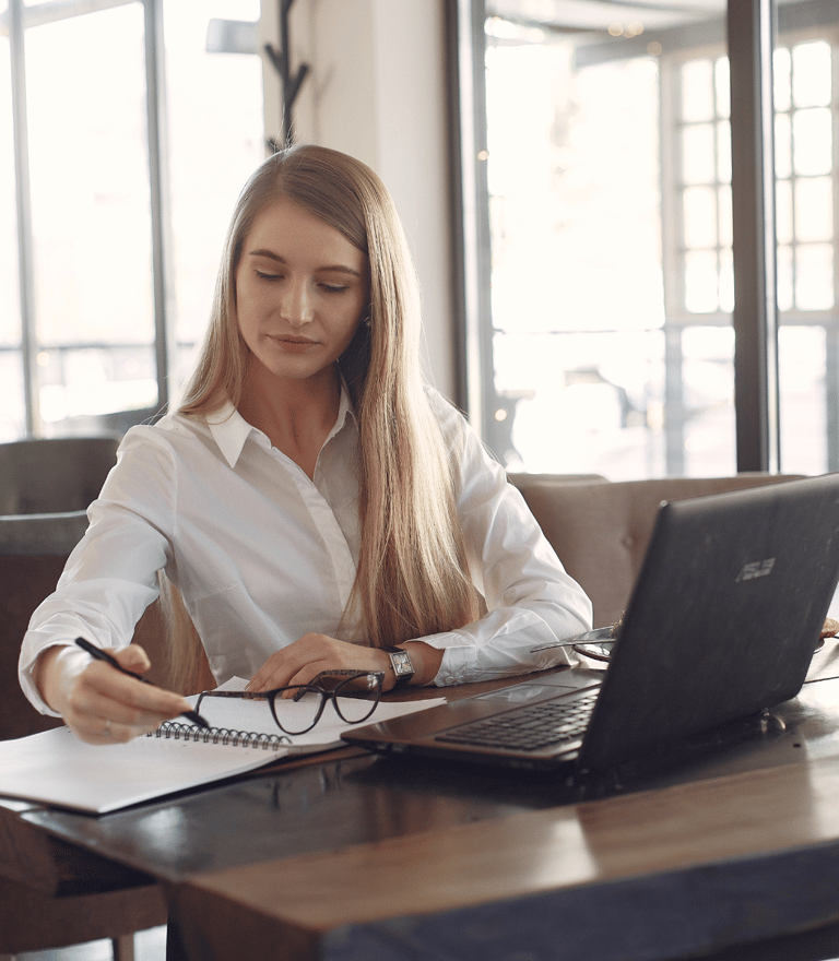 a woman sitting at a table with a laptop, notebook, pen and glasses