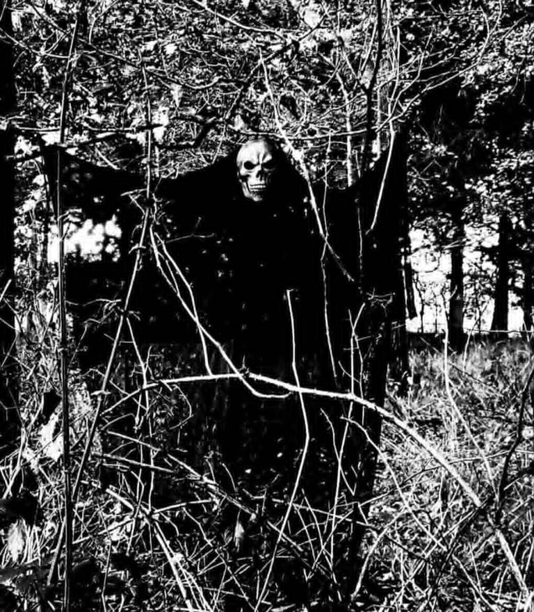 Black and white photo of a hooded and robed skeleton lurking in dense undergrowth