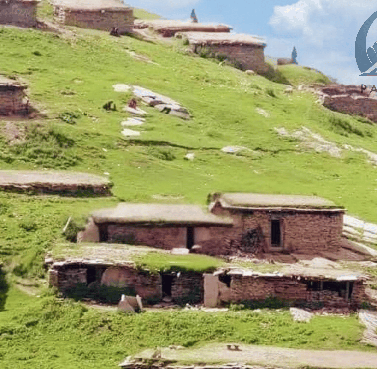 a mountain scene with a cow grazing in the grass along with old mud houses