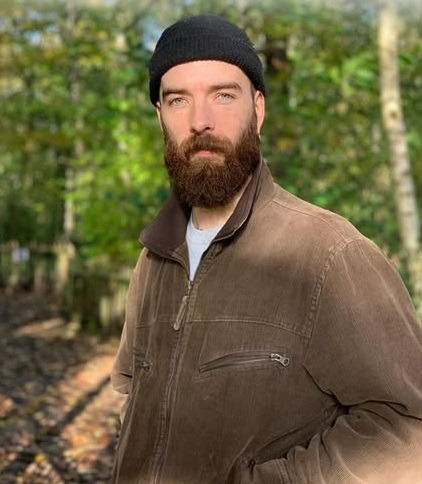 Bearded man wearing a black beanie and brown jacket posing outdoors in a sunny forest.