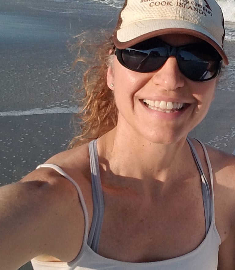 A smiling woman wearing sunglasses and a baseball hat takes a selfie on a sunny beach.
