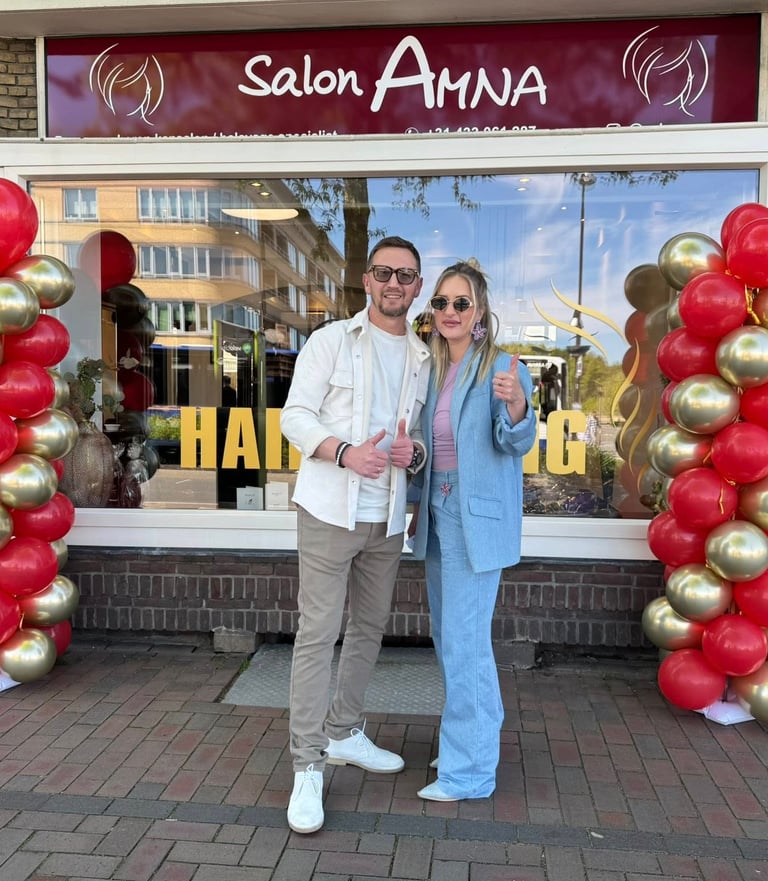 A couple posing in front of Salon Amna hair salon entrance decorated with red and gold balloon arches.