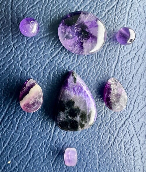 Amatista y charoíta pulidas en diferentes formas sobre una superficie azul texturizada.