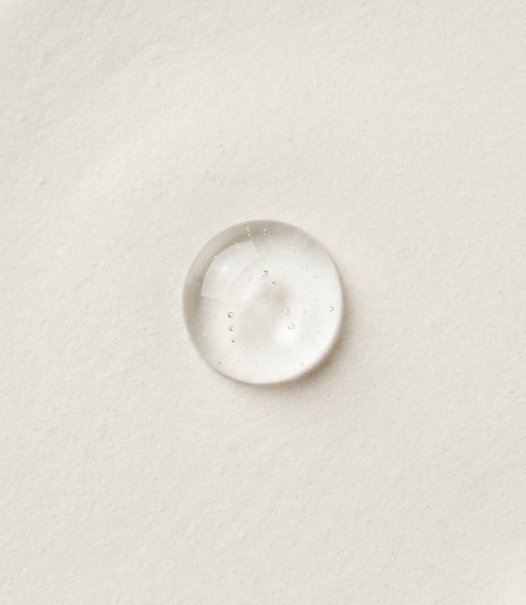 Overhead macro photography of a single clear cosmetic gel droplet on a warm off-white surface, bright diffused clinical studio light, sharp focus on texture