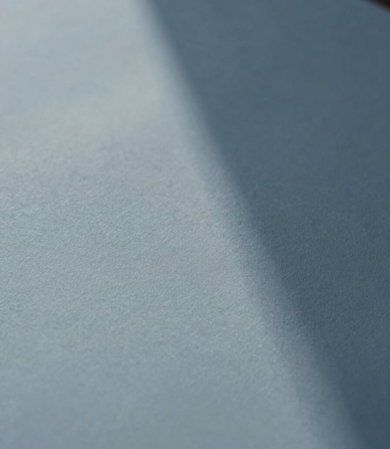An extreme close-up of fine textured paper casting a soft geometric shadow under diffused daylight, minimalist editorial style, muted blue tones, shallow depth of field.