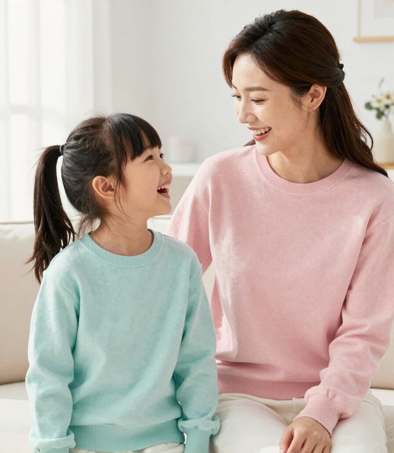 A smiling mother and young daughter wearing coordinated pastel cotton sweaters, laughing together in a bright, sunlit living room, high-key natural light, 35mm lens