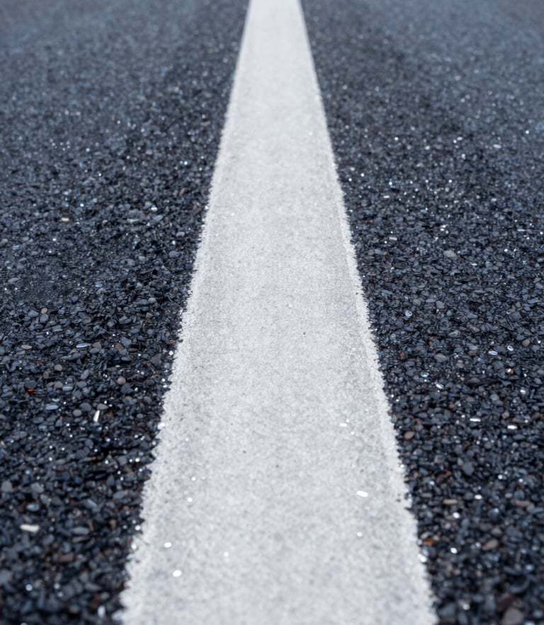 Low-angle perspective shot looking down a perfectly straight, wet white thermoplastic road line on dark asphalt, sharp glass beads reflecting morning light.