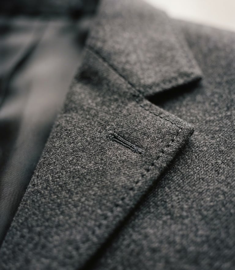 Macro studio photograph of a structured charcoal suit jacket lapel, soft natural window light, highlighting premium wool texture and clean hand-finished stitching, 35mm
